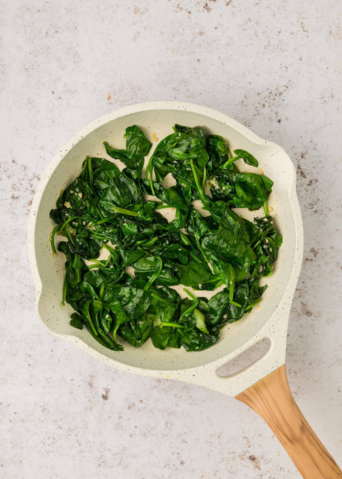 A white frying pan with sautéed spinach on a light, textured surface.