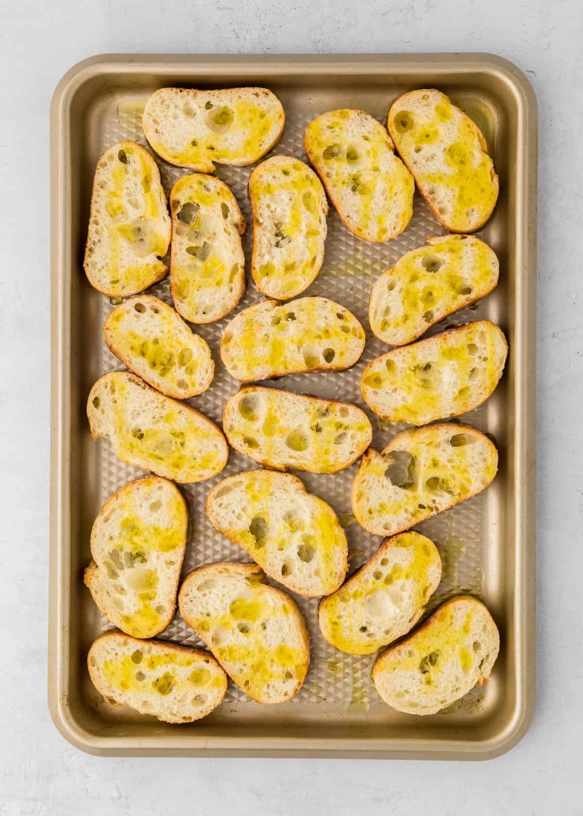 Slices of baguette drizzled with olive oil on a baking sheet, ready to be toasted.