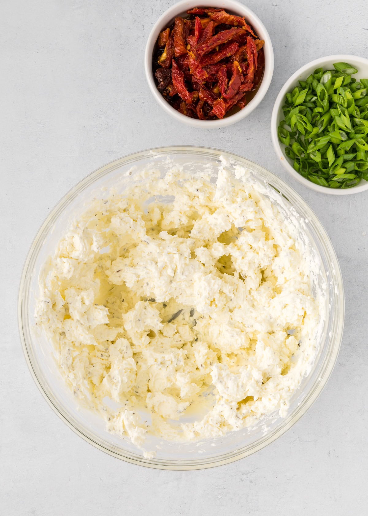 A bowl of whipped cheese with bowls of chopped green onions and sun-dried tomatoes nearby.
