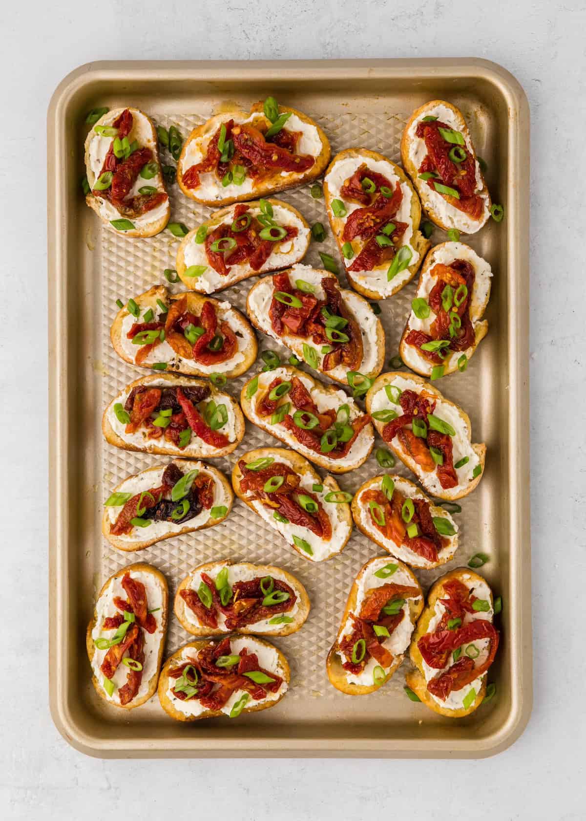Toasted bread slices with cheese, sun-dried tomatoes, and green onions on a baking tray.