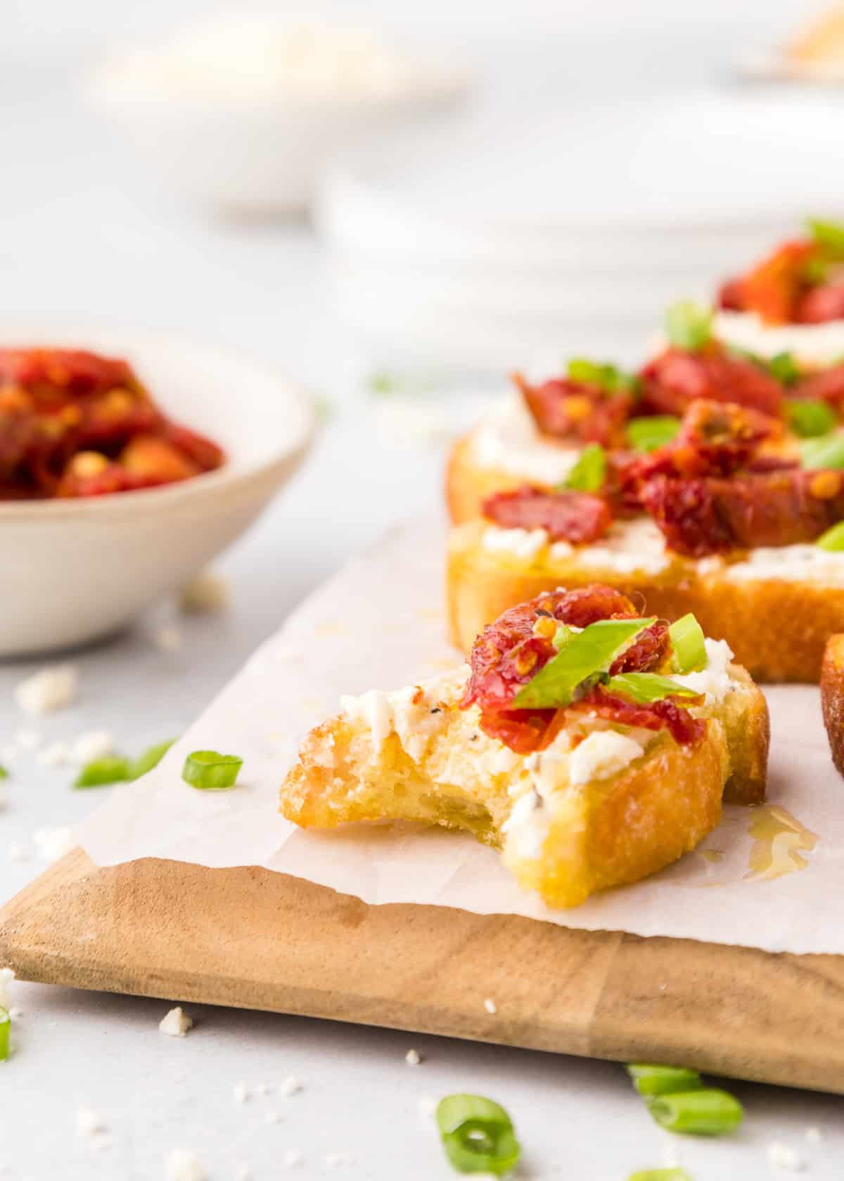 Crostini with cheese, sun-dried tomatoes, and green onions on parchment, one with a bite.