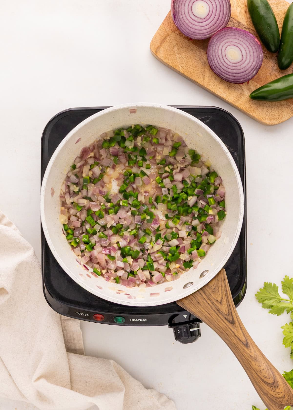 Diced onions and jalapeños sautéing in a pan on a stovetop, with veggies nearby.