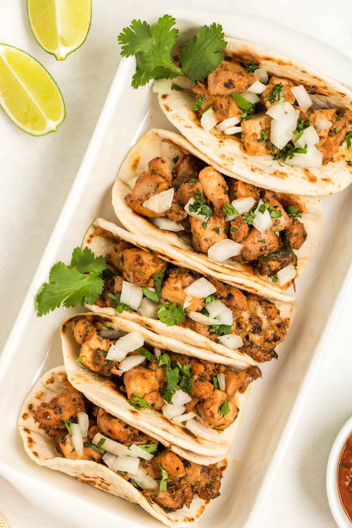 Four chicken tacos with onions and cilantro on a white tray, with lime wedges on the side.