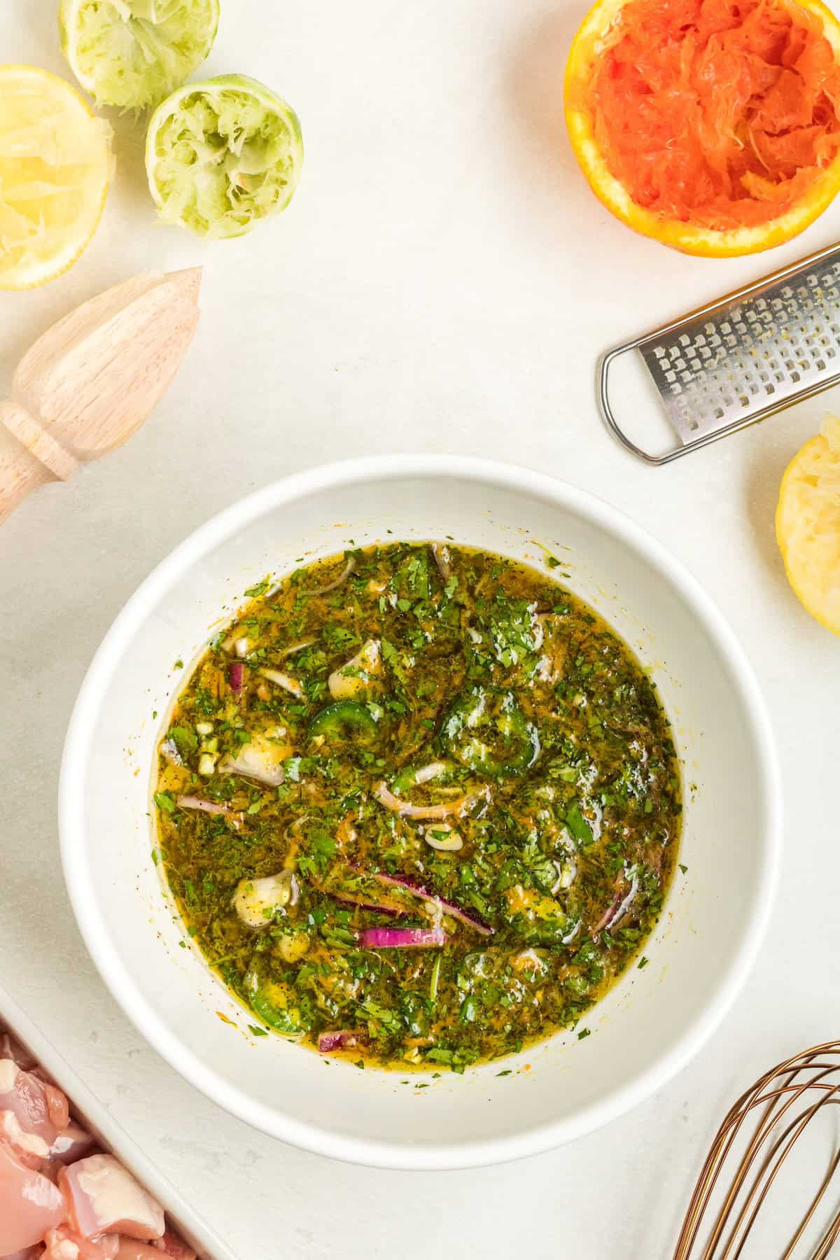 Bowl of citrus herb marinade with citrus fruits and kitchen tools on a white surface.