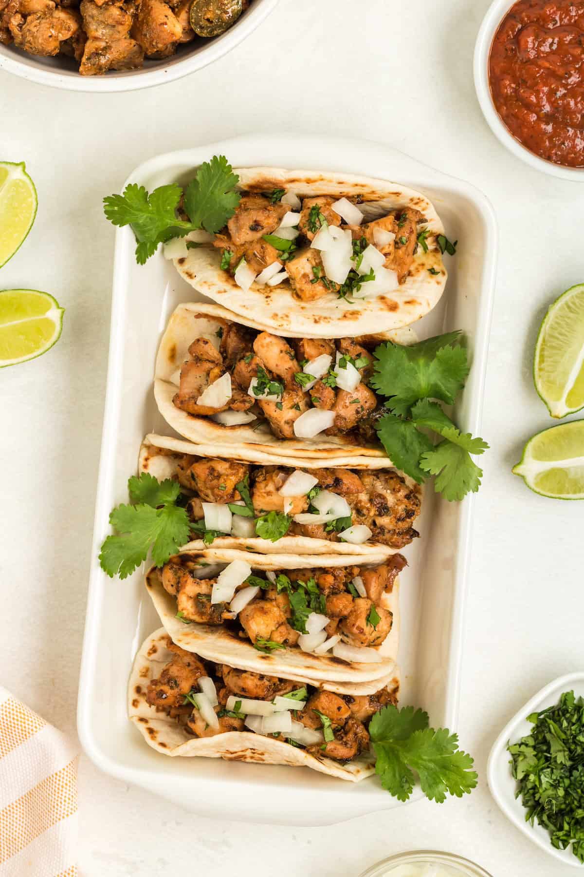 Four chicken tacos topped with onions and cilantro, served with lime wedges and salsa.