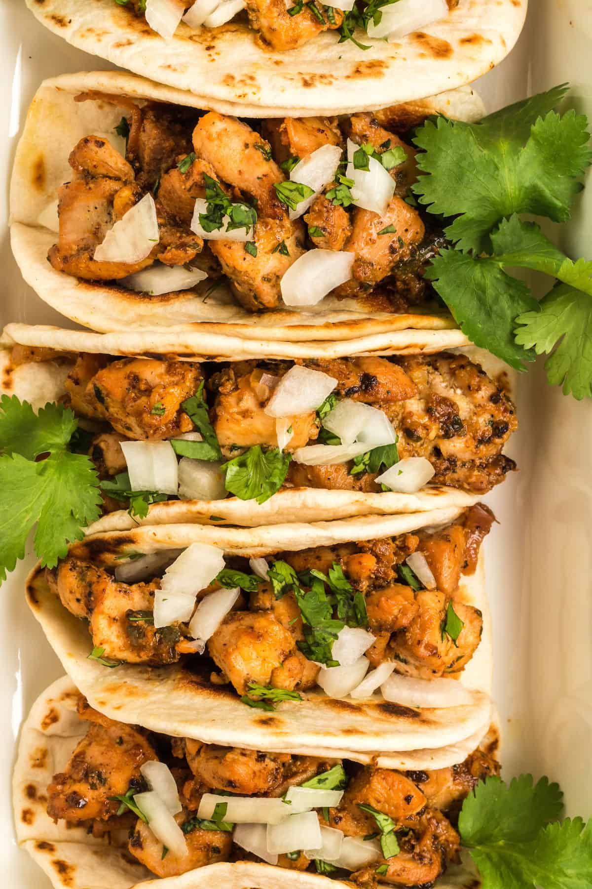 Four chicken tacos topped with chopped onion and cilantro, garnished with fresh cilantro leaves.