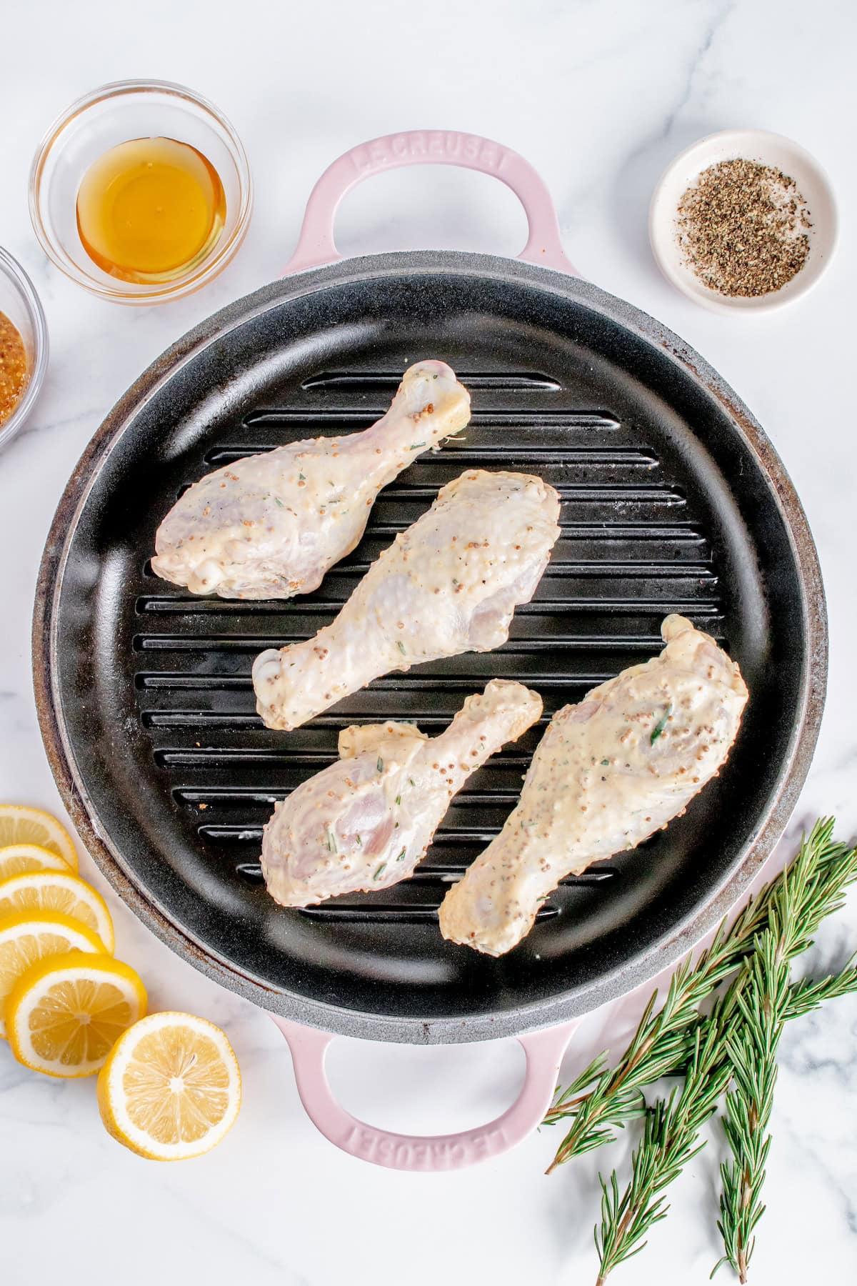 Raw seasoned chicken drumsticks on a grill pan, with lemon slices, rosemary, and spices nearby.