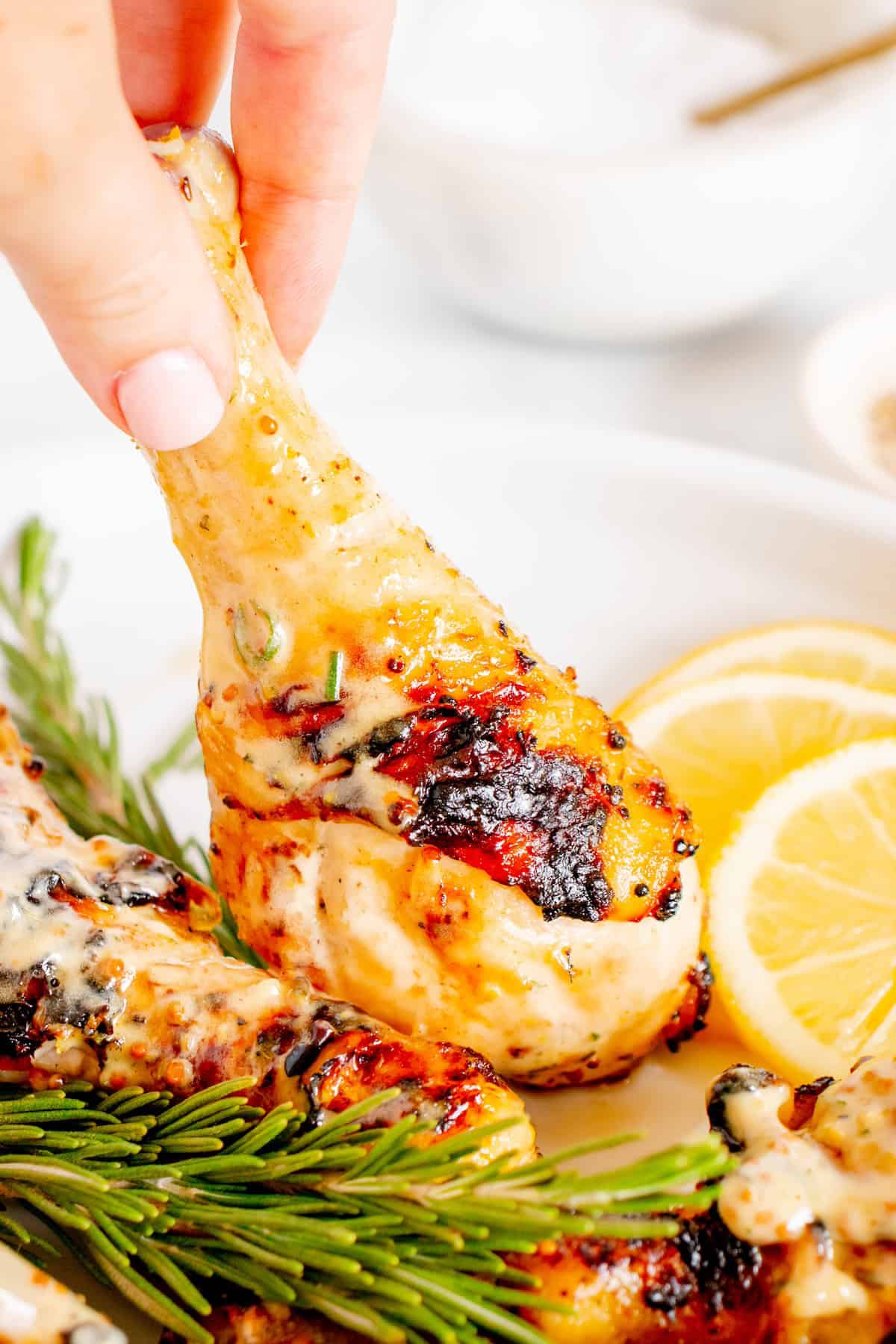 A hand holding a grilled chicken drumstick garnished with herbs and lemon slices.