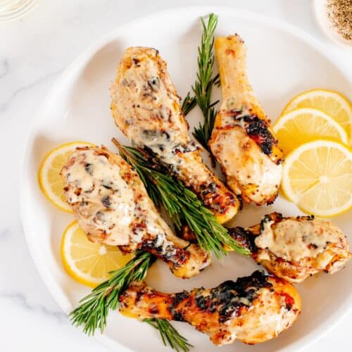 Grilled chicken drumsticks with creamy sauce, lemon slices, and rosemary on a white plate.