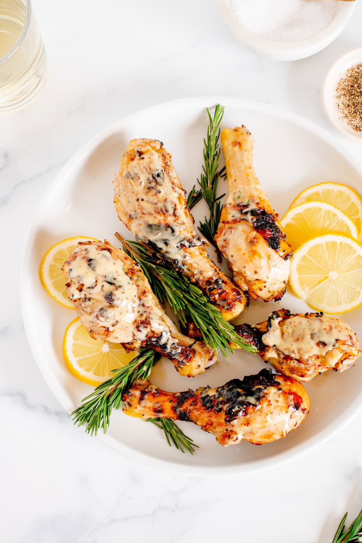 Grilled chicken drumsticks with creamy sauce, lemon slices, and rosemary on a white plate.