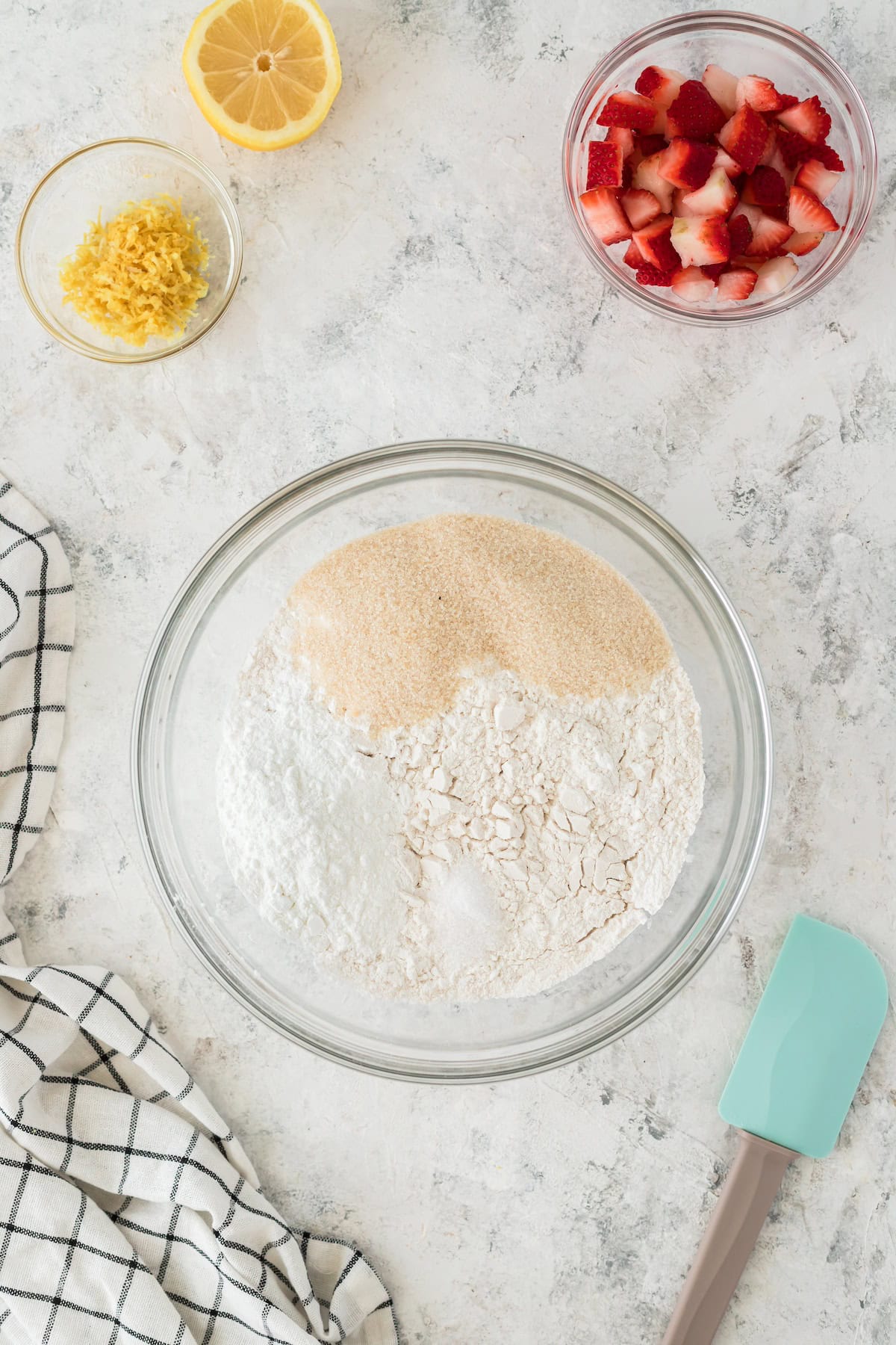 Large bowl with flour and sugar, lemon zest, lemon, chopped strawberries, towel, and spatula.