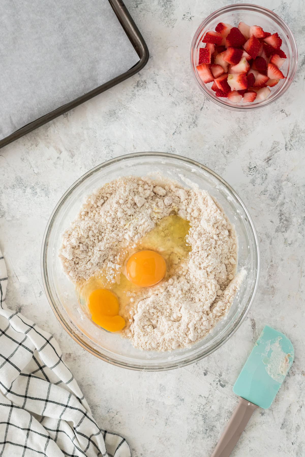 A bowl with flour and two eggs, chopped strawberries, spatula, and baking tray nearby.