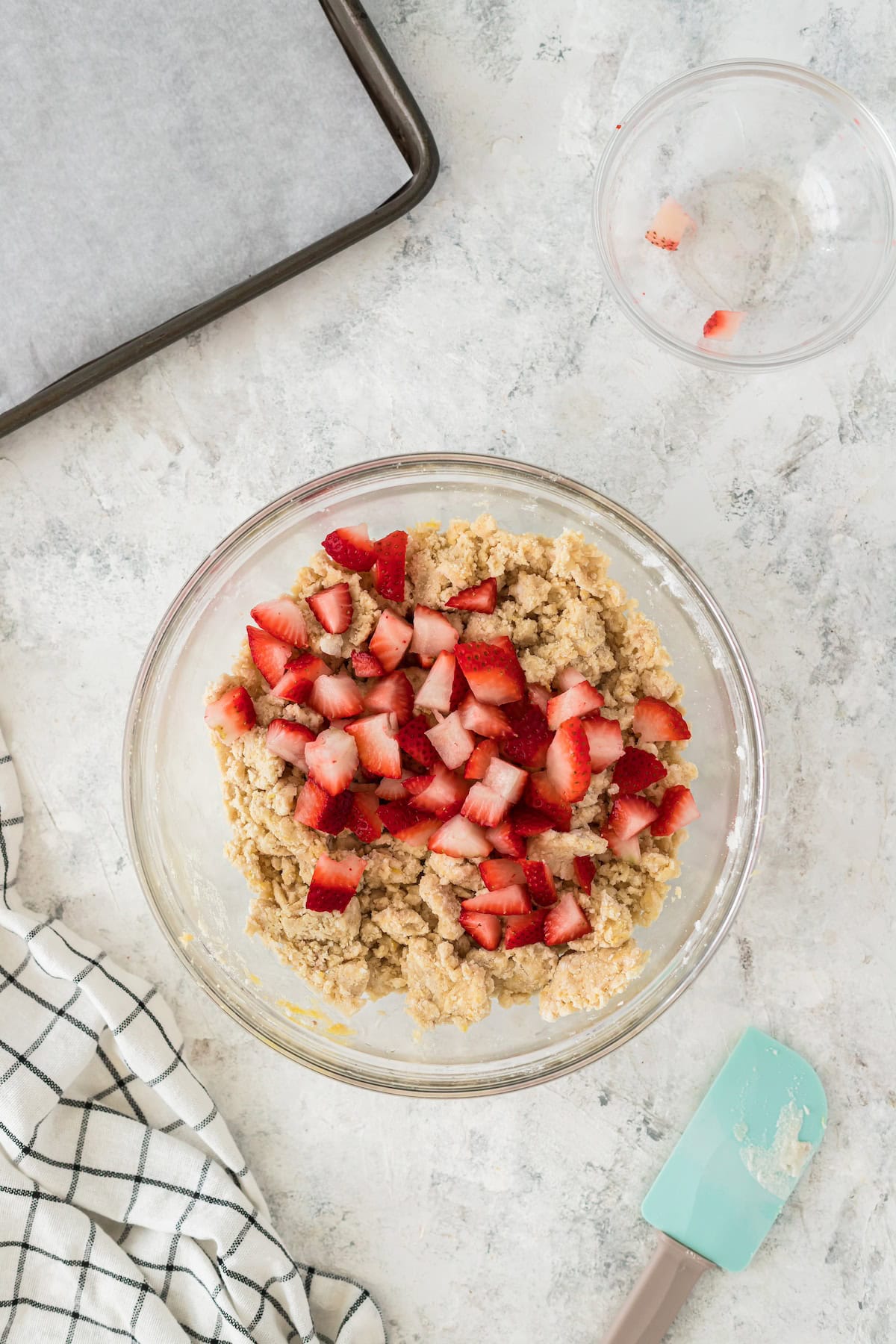 Glass bowl with dough and chopped strawberries, next to a spatula, towel, and baking tray.