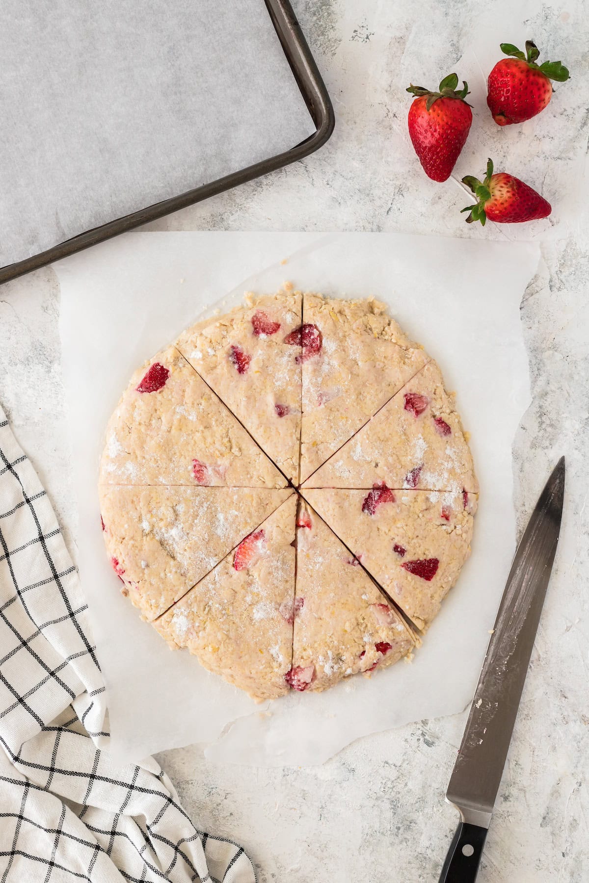 Round strawberry scone dough cut into wedges on parchment, with knife and strawberries nearby.