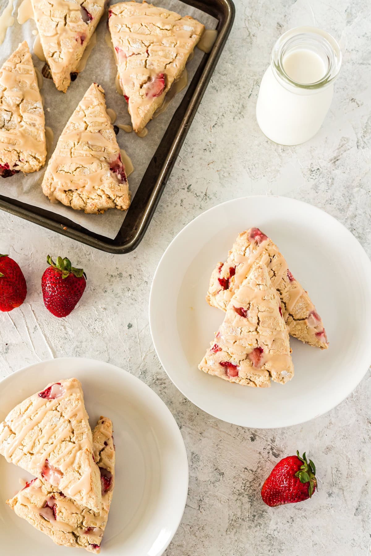 Strawberry scones on plates and a baking tray with fresh strawberries and a bottle of milk.