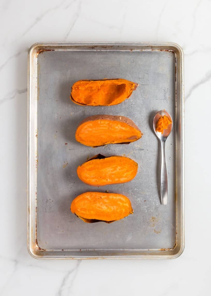 Four baked sweet potato halves and a spoon on a metal baking sheet, shot from above.