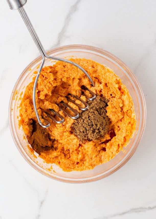 A bowl of mashed sweet potatoes with brown sugar and a masher on a white surface.