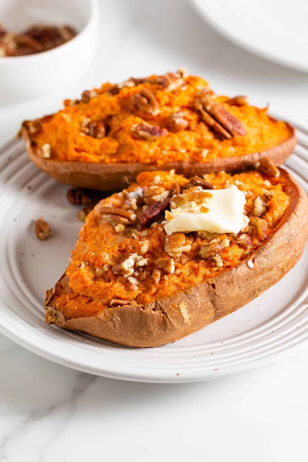Two baked sweet potato halves topped with pecans and a pat of butter on a white plate.