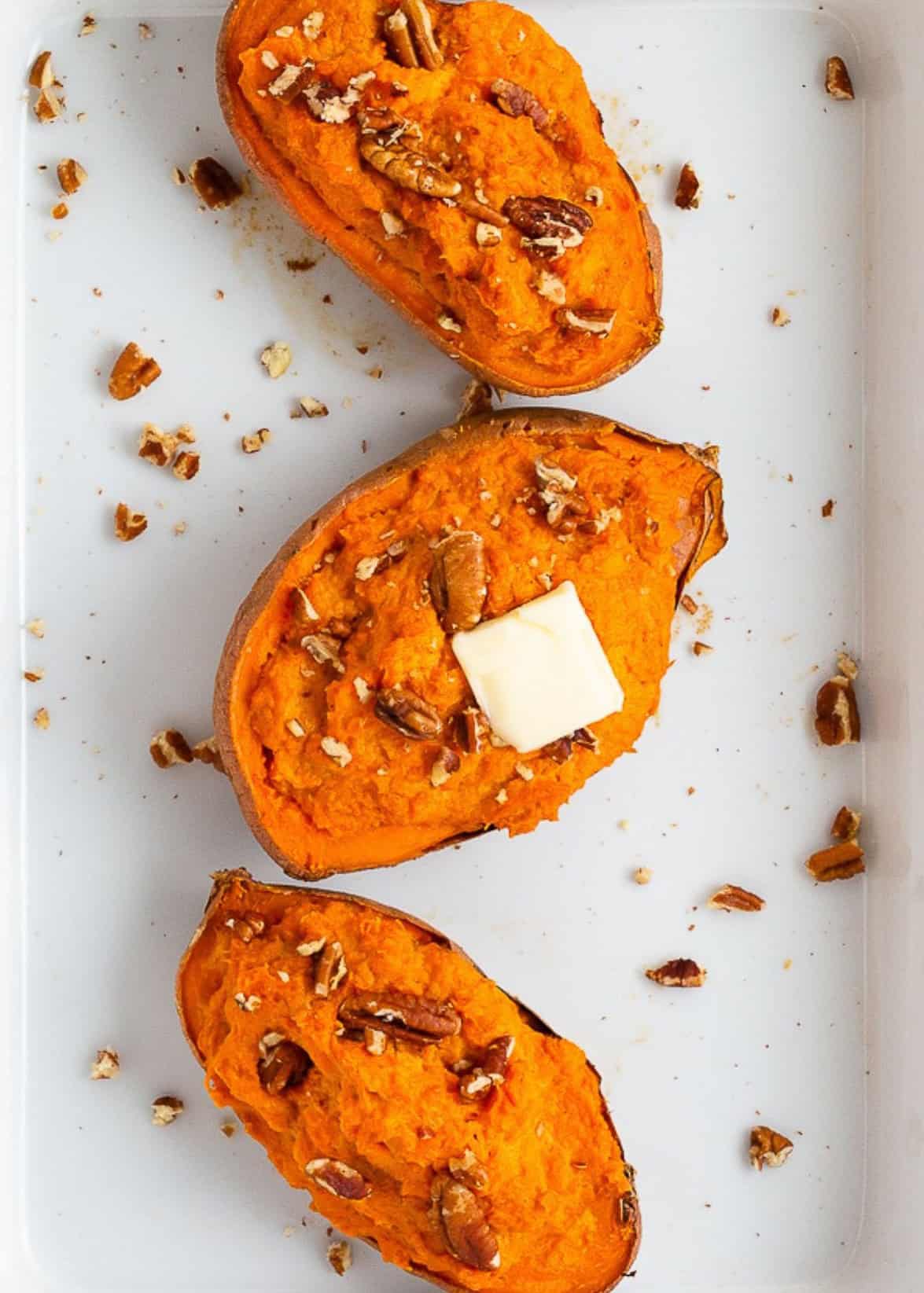 Three baked sweet potato halves topped with pecans and a pat of butter on a white surface.