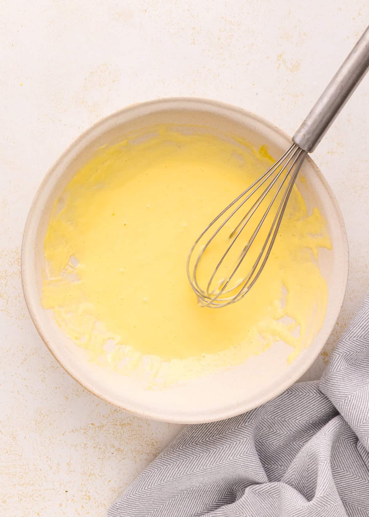 A whisk in a bowl of yellow batter for Air Fryer Chicken Tenders beside a striped cloth.