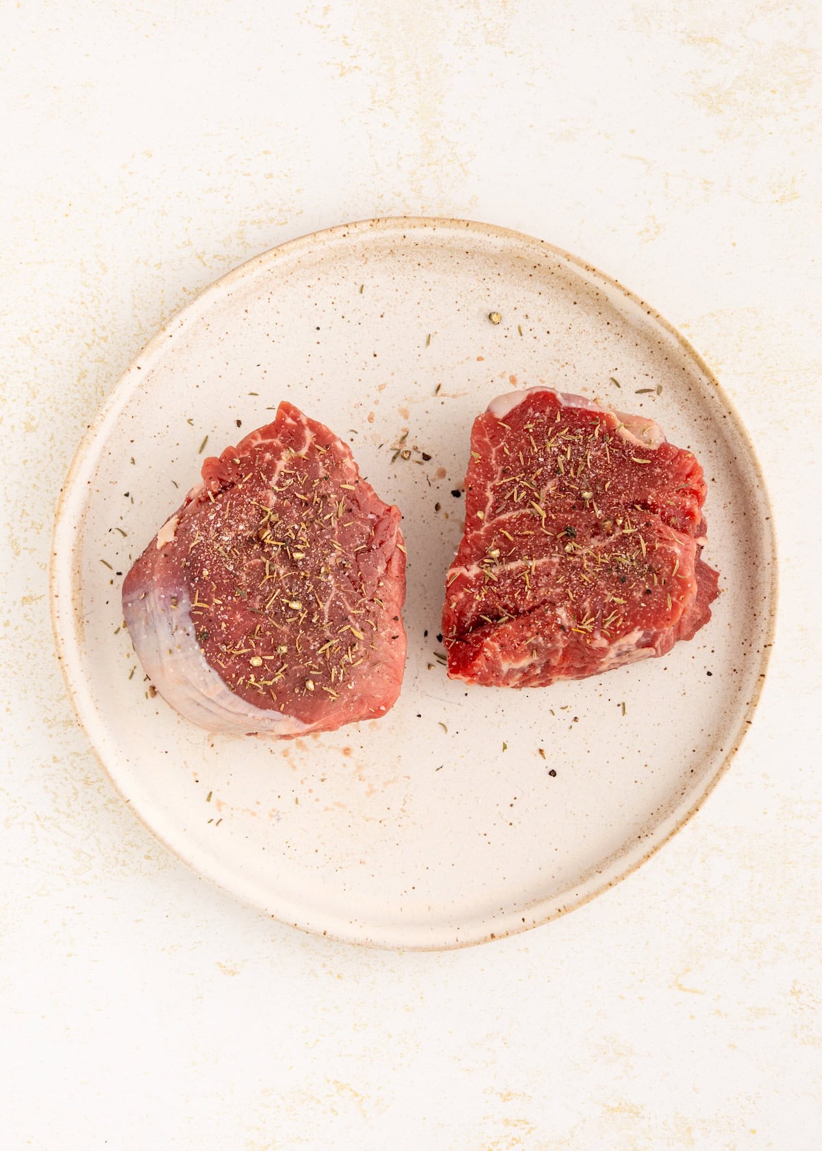 Two raw filet mignon steaks with seasoning on a beige ceramic plate.