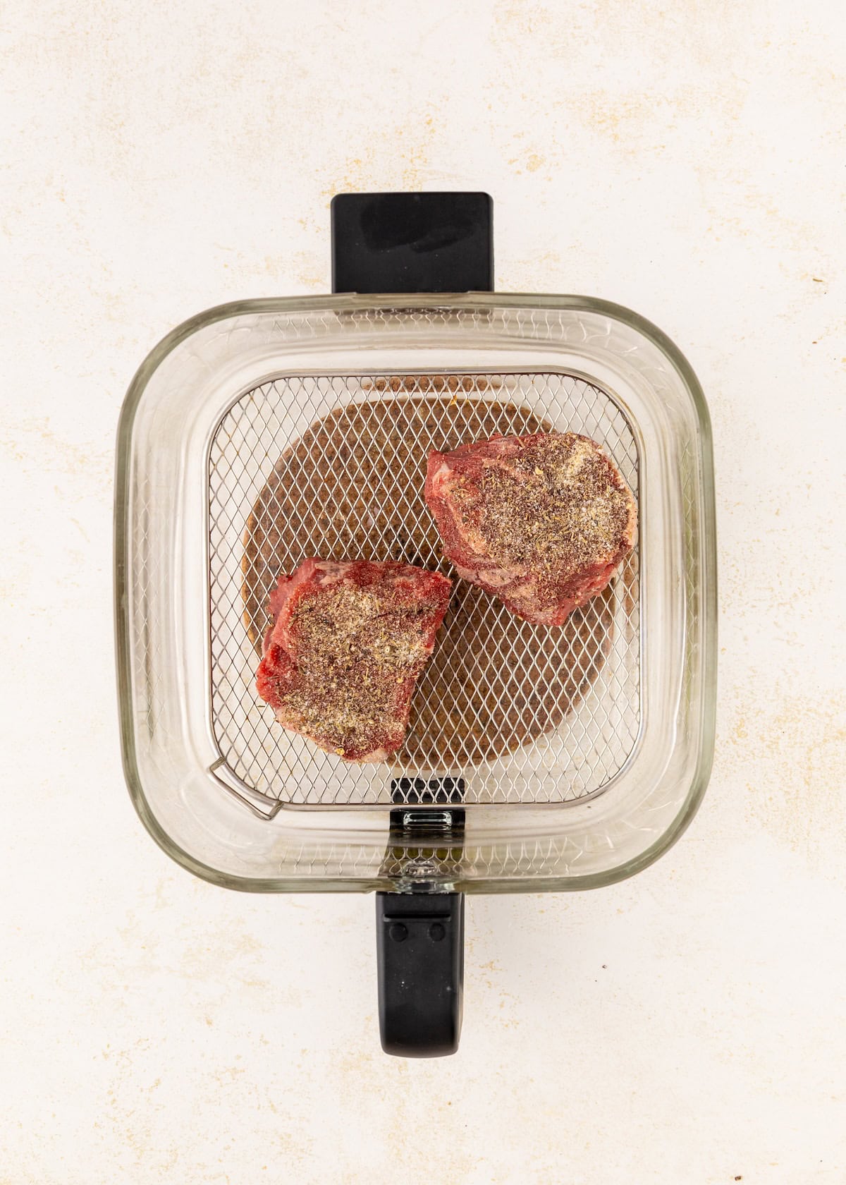 Two raw, seasoned steaks in an air fryer basket on a light background.
