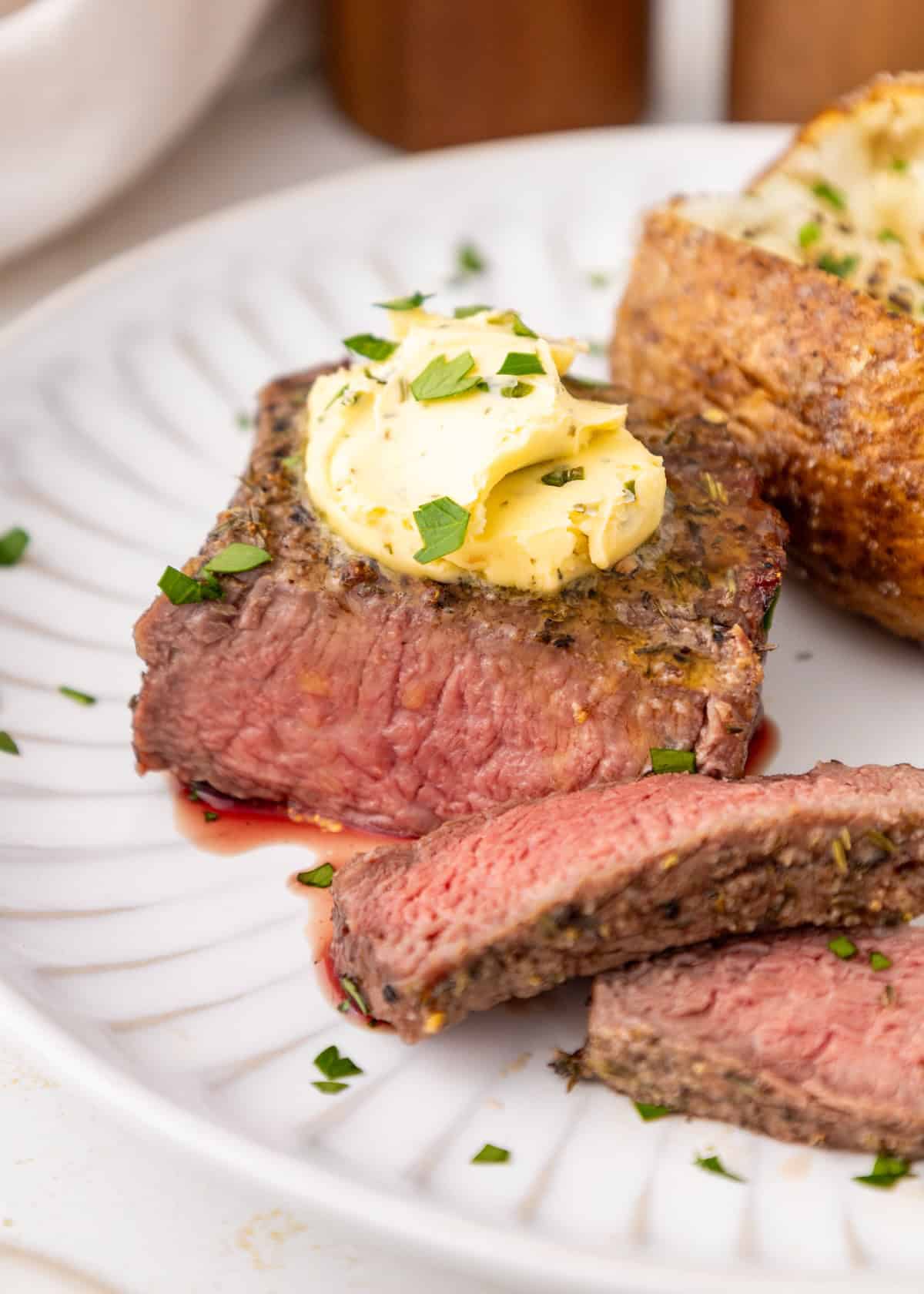 A medium-rare steak with herb butter and parsley, served with a baked potato on a white plate.