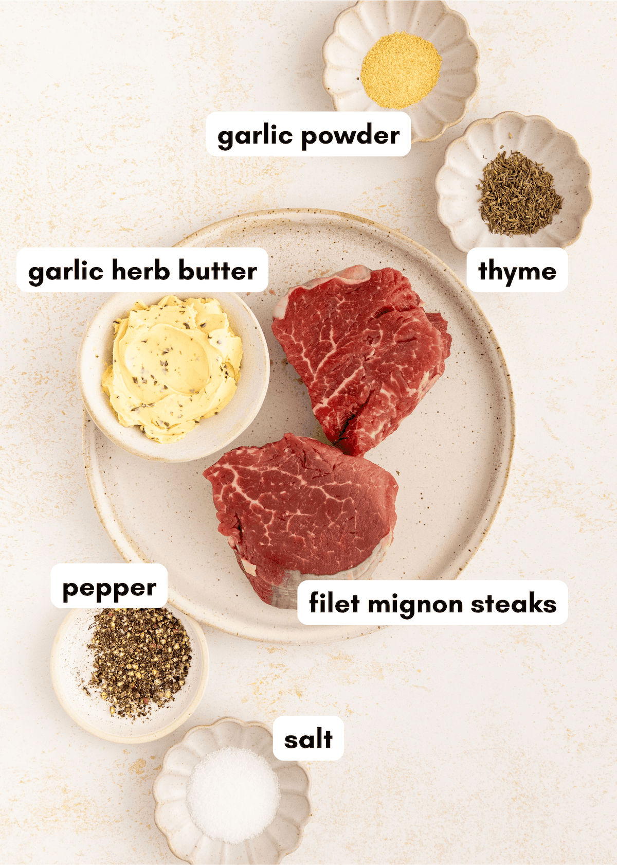 Two raw filet mignon steaks with garlic herb butter, spices, salt, pepper, and thyme in bowls.
