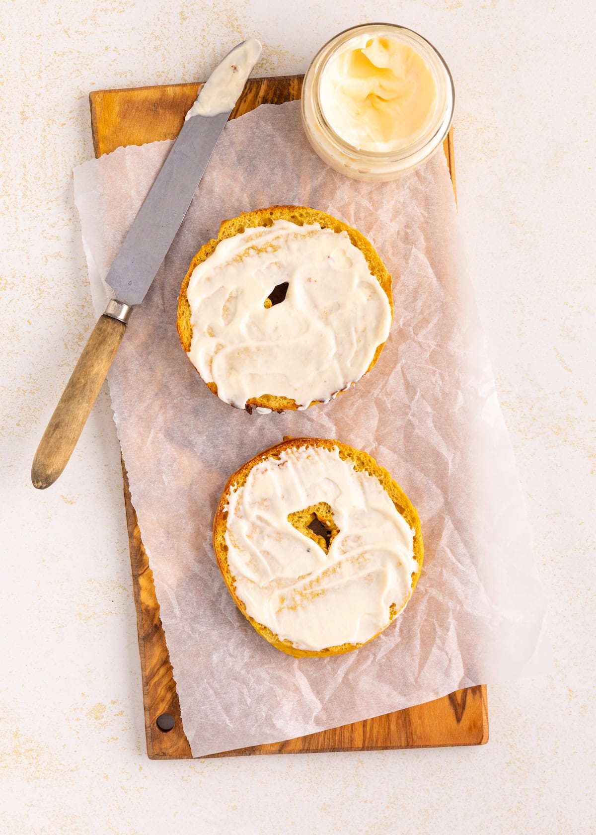 A sliced bagel with cream cheese, knife, and jar on parchment paper and wooden board.