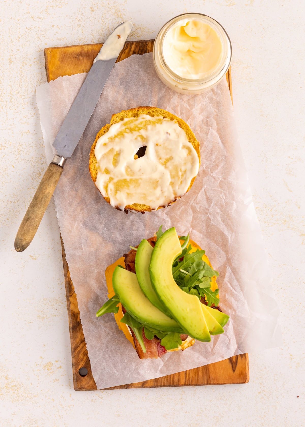 Open sandwich with avocado, bacon, cheese, arugula, and mayo on parchment paper and a board.