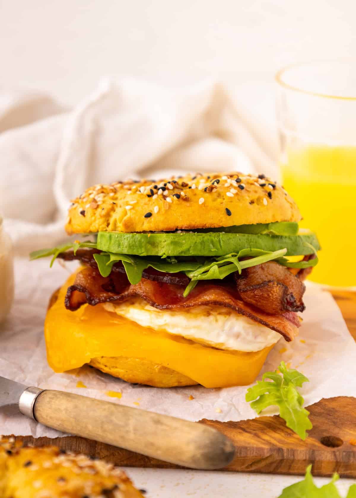 A breakfast sandwich with bacon, egg, cheese, avocado, and greens on a seeded bun.