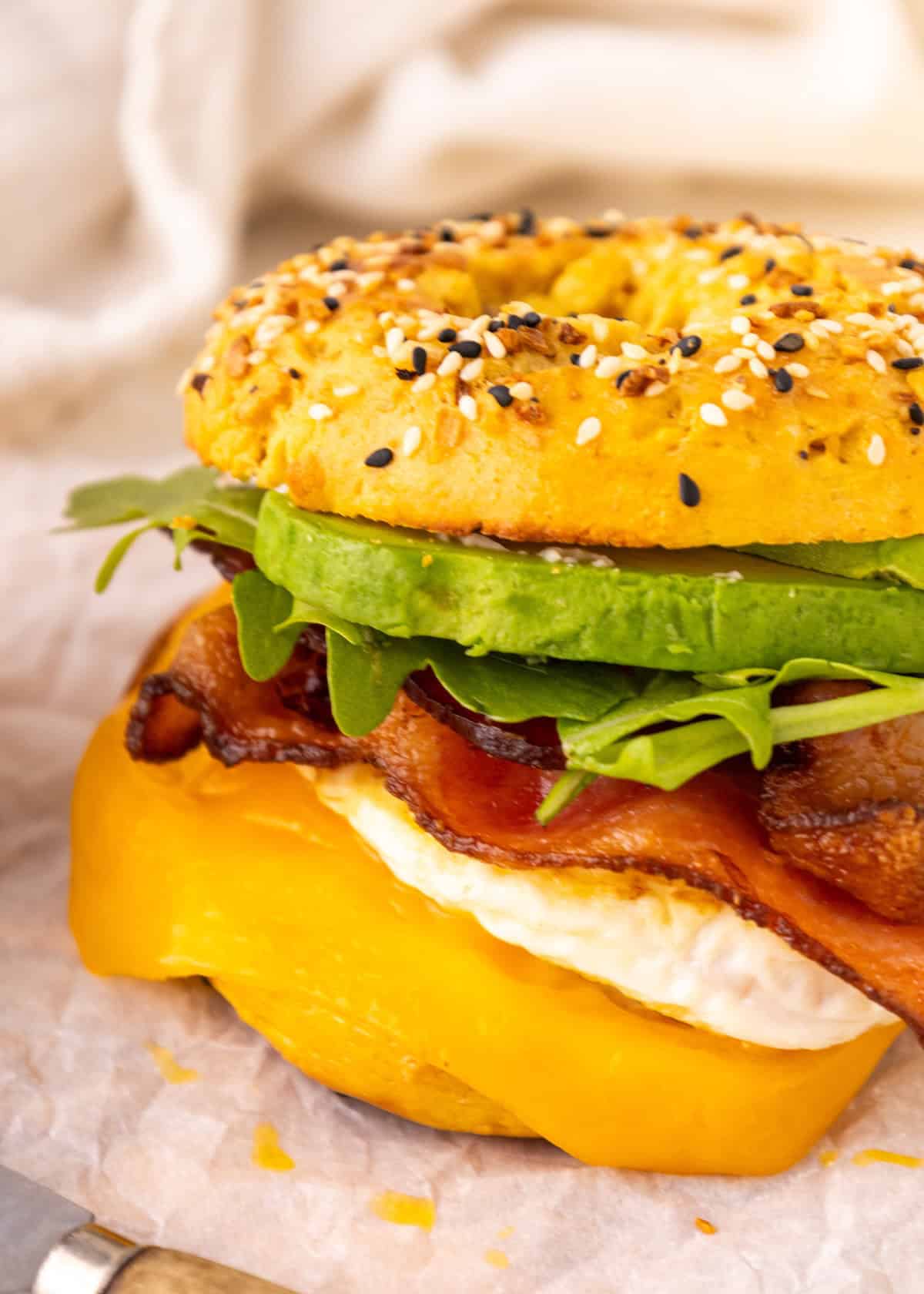 Everything bagel sandwich with egg, bacon, avocado, arugula, and cheddar cheese on parchment.