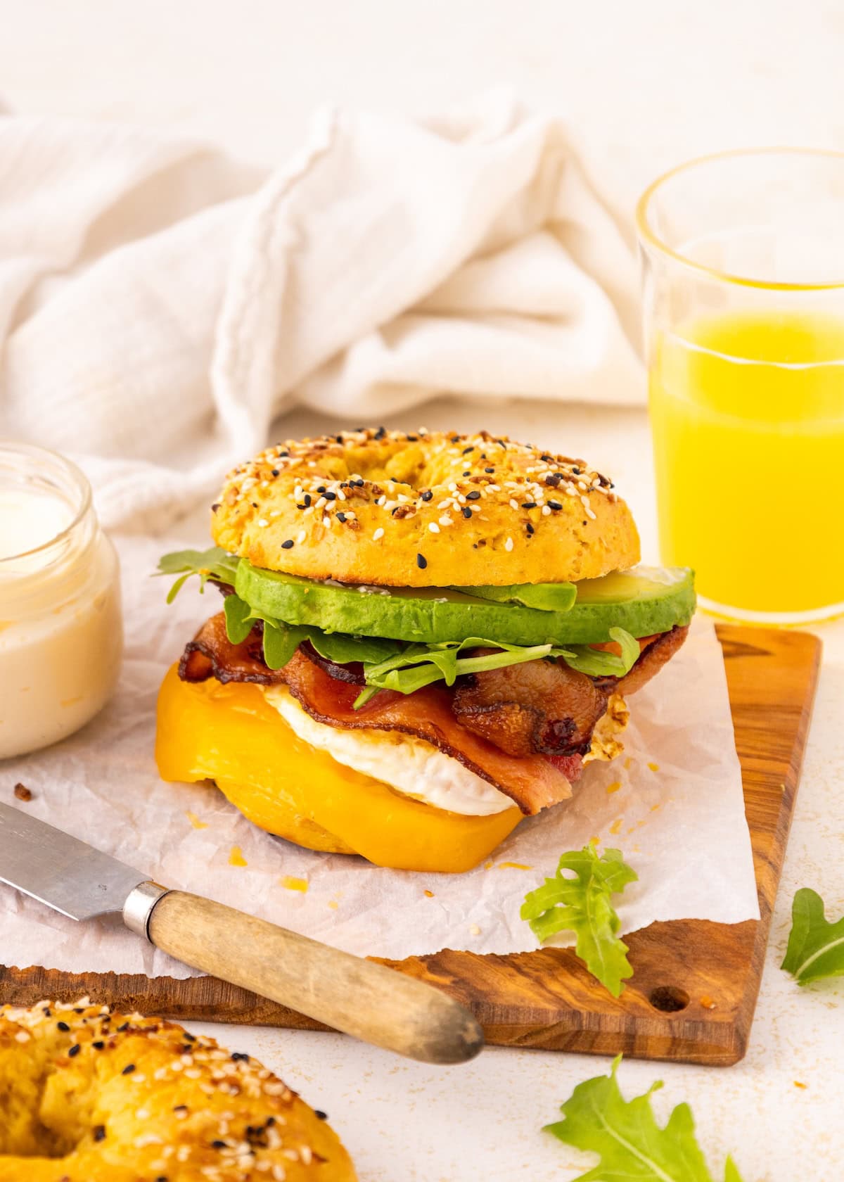 Bagel sandwich with bacon, cheese, avocado, arugula, sauce, and a glass of orange juice.