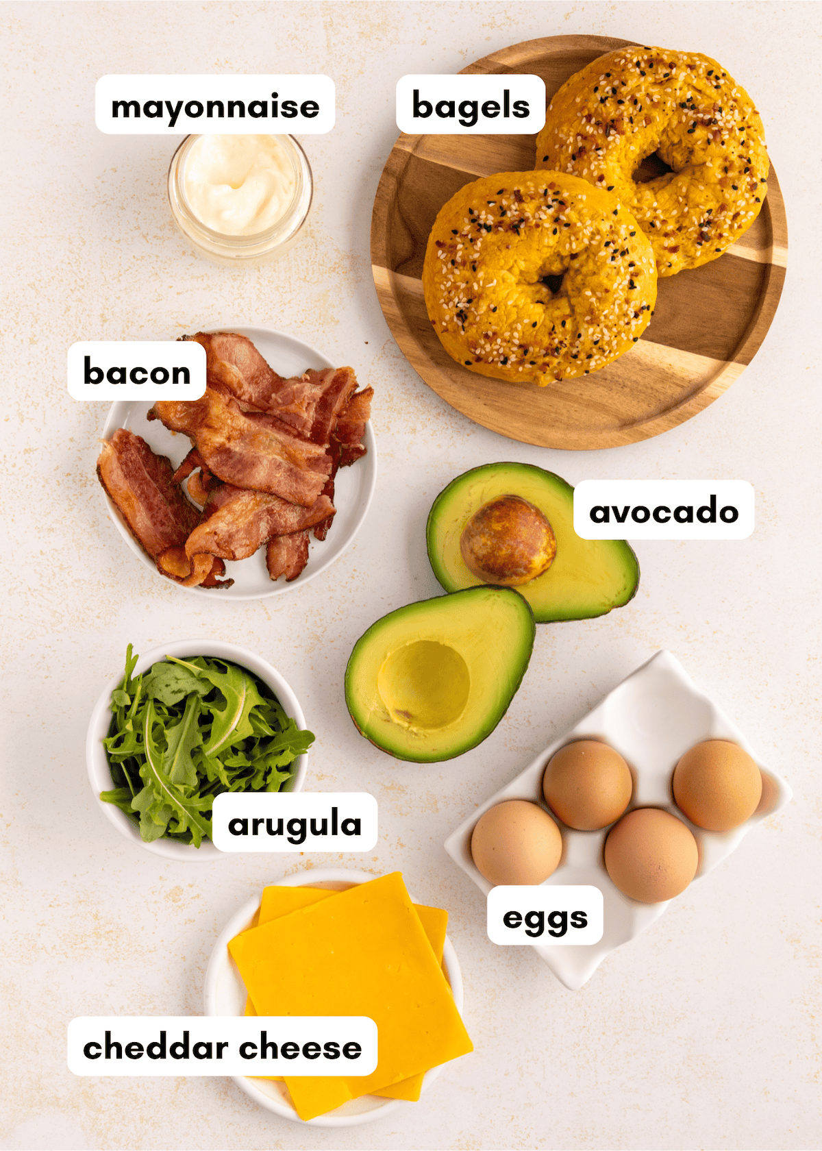 Bagels, mayonnaise, bacon, avocado, arugula, eggs, and cheddar cheese on a light surface.