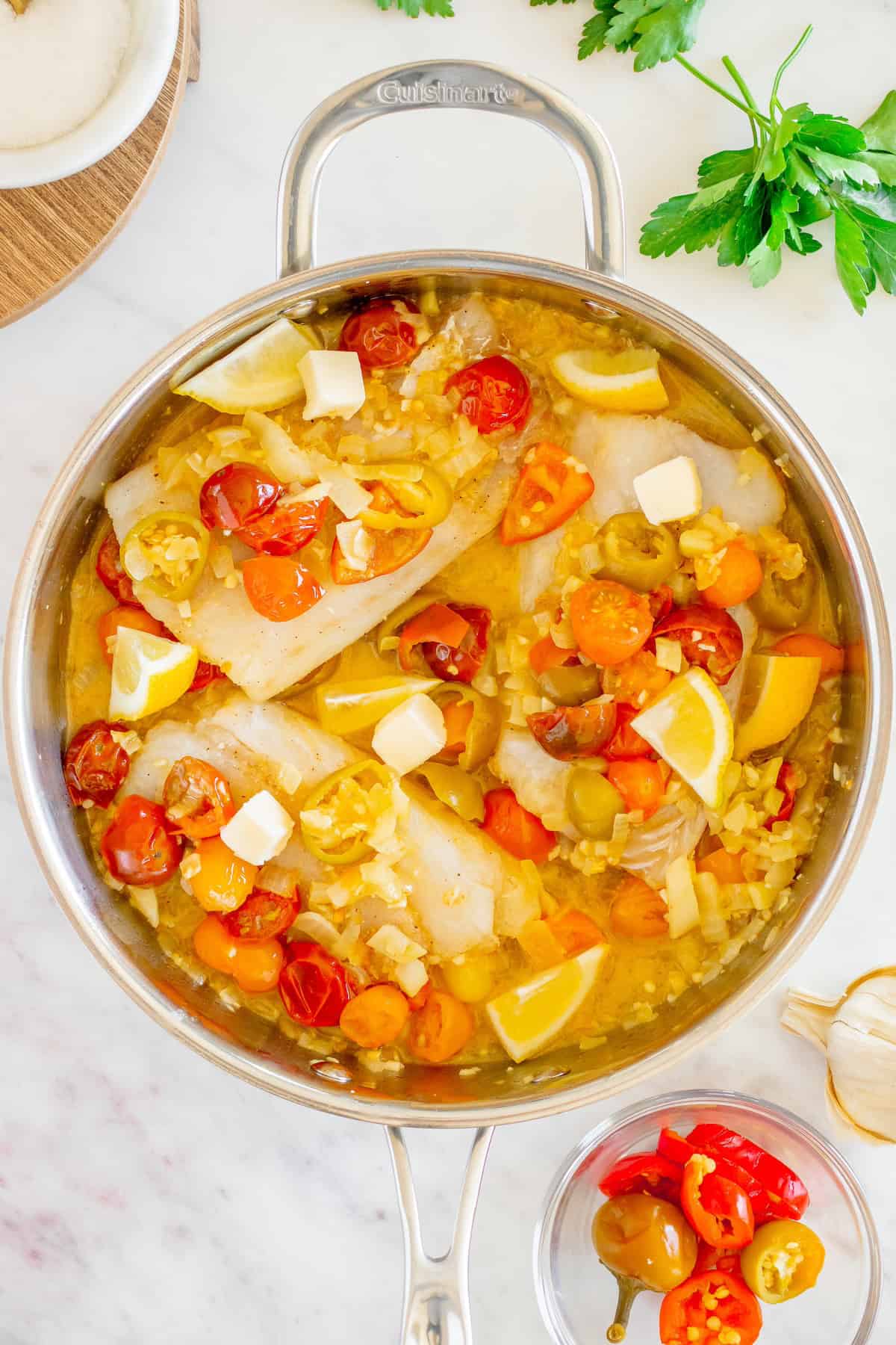 Fish fillets in a pan with cherry tomatoes, lemon wedges, herbs, and butter on a white surface.