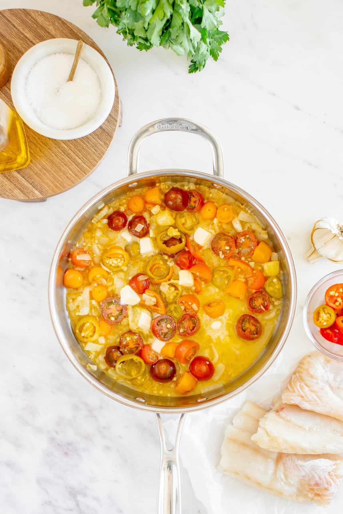 Sliced cherry tomatoes cooking in a pan with garlic and oil, cod fillets and seasonings nearby.