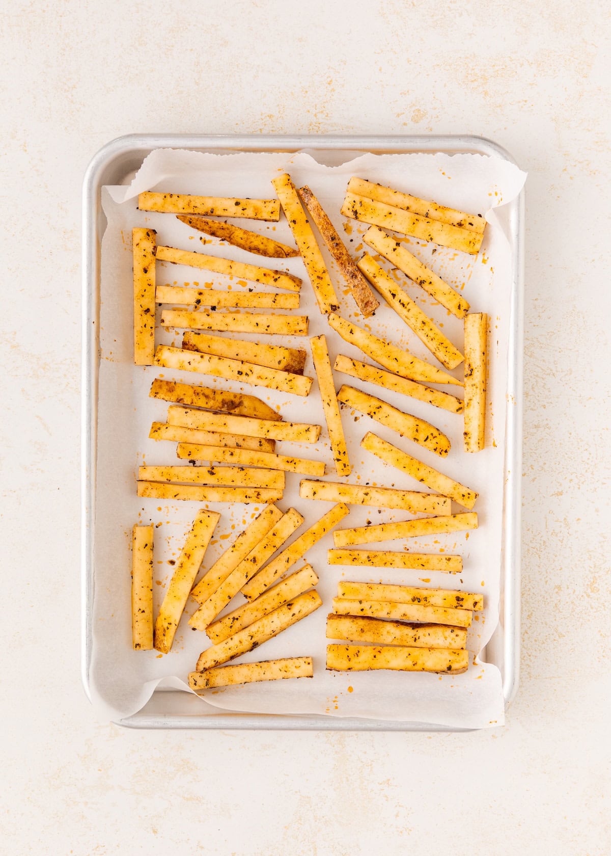 Seasoned fries spread on a parchment-lined baking tray, ready to be baked.