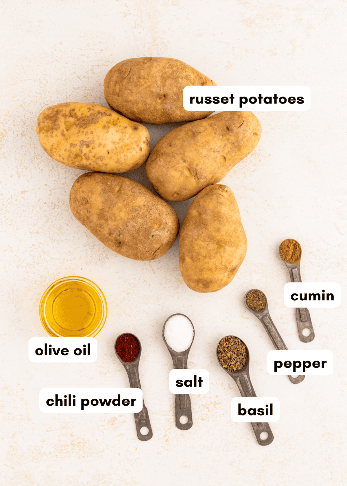 Six russet potatoes, olive oil, and labeled spices on a light surface.