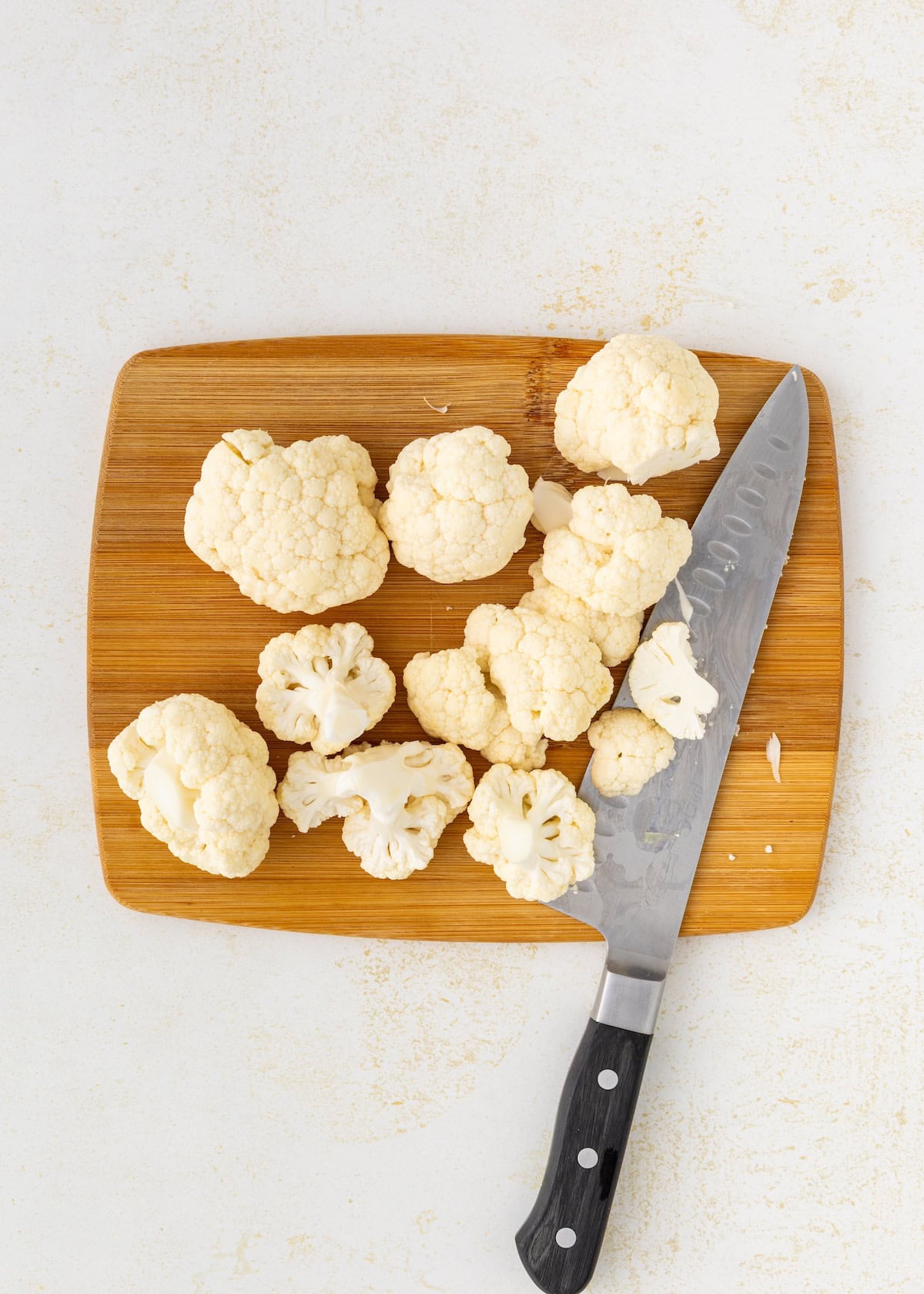 Chopped cauliflower for Buffalo Cauliflower Wings sits with a knife on a wooden cutting board.