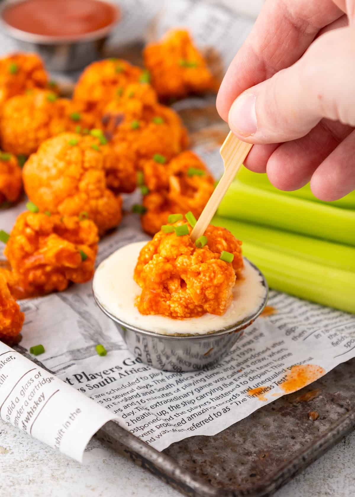 A hand dips Buffalo Cauliflower Wings into creamy sauce next to celery sticks on newspaper.
