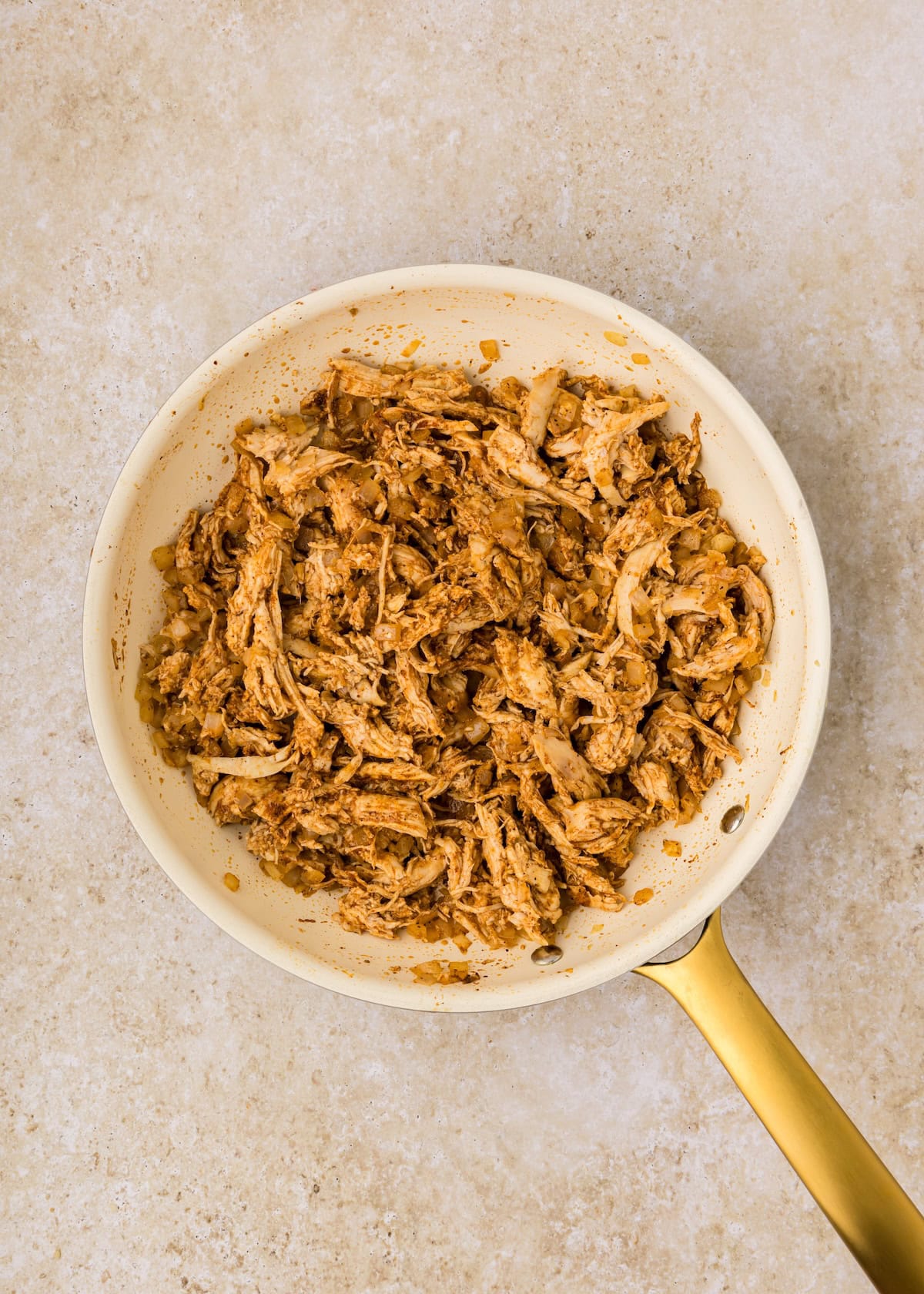 Shredded seasoned chicken cooking in a white skillet with a gold handle on a light surface.