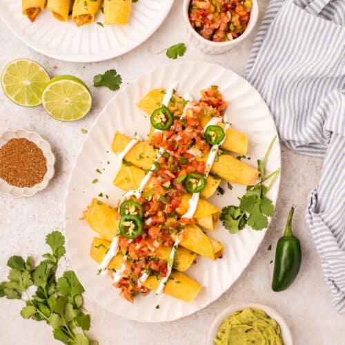 Plates of taquitos topped with salsa, jalapeños, and sour cream, with sides of guacamole and limes.