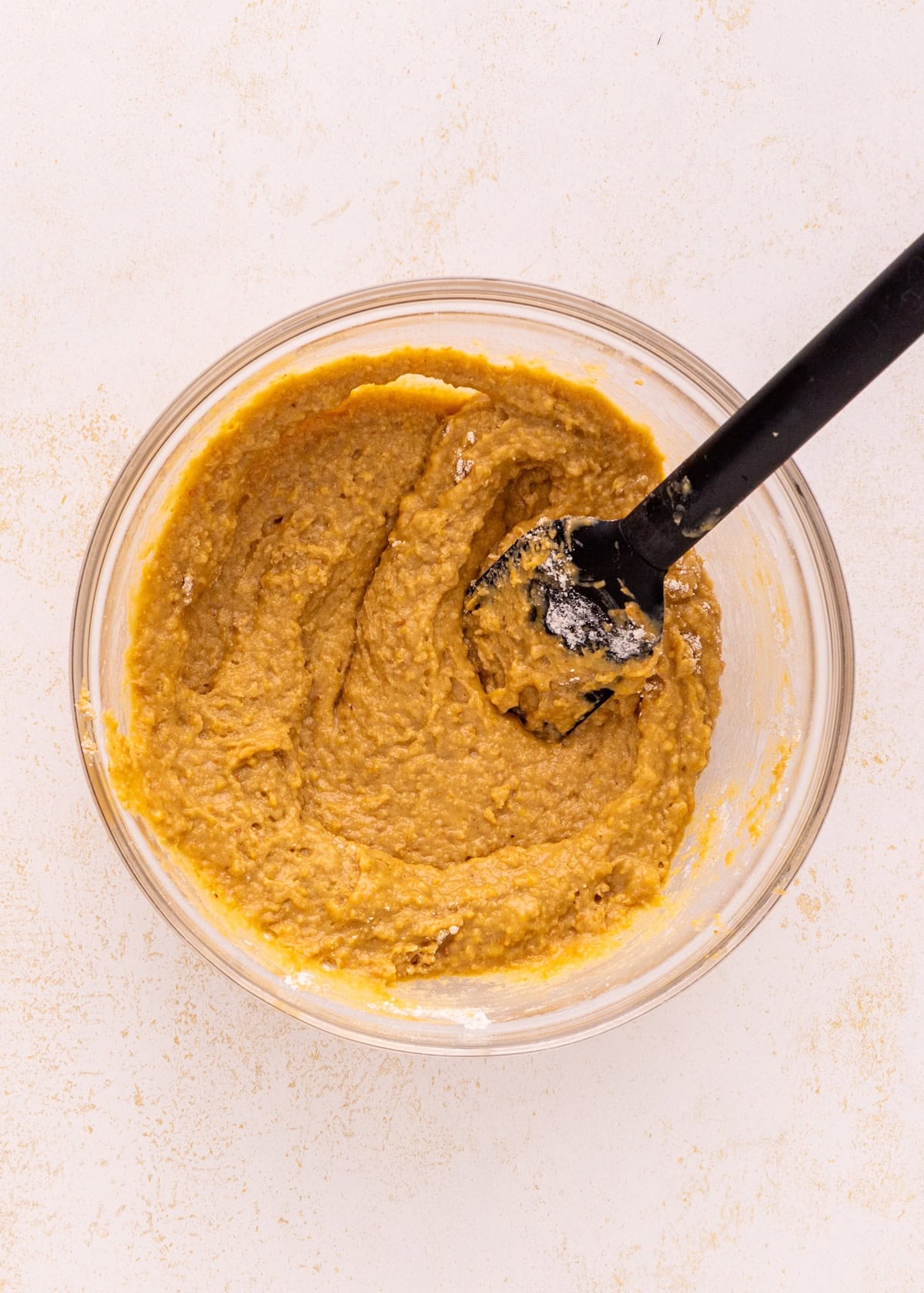 Bowl of thick, light brown batter with a black spatula on a light surface.