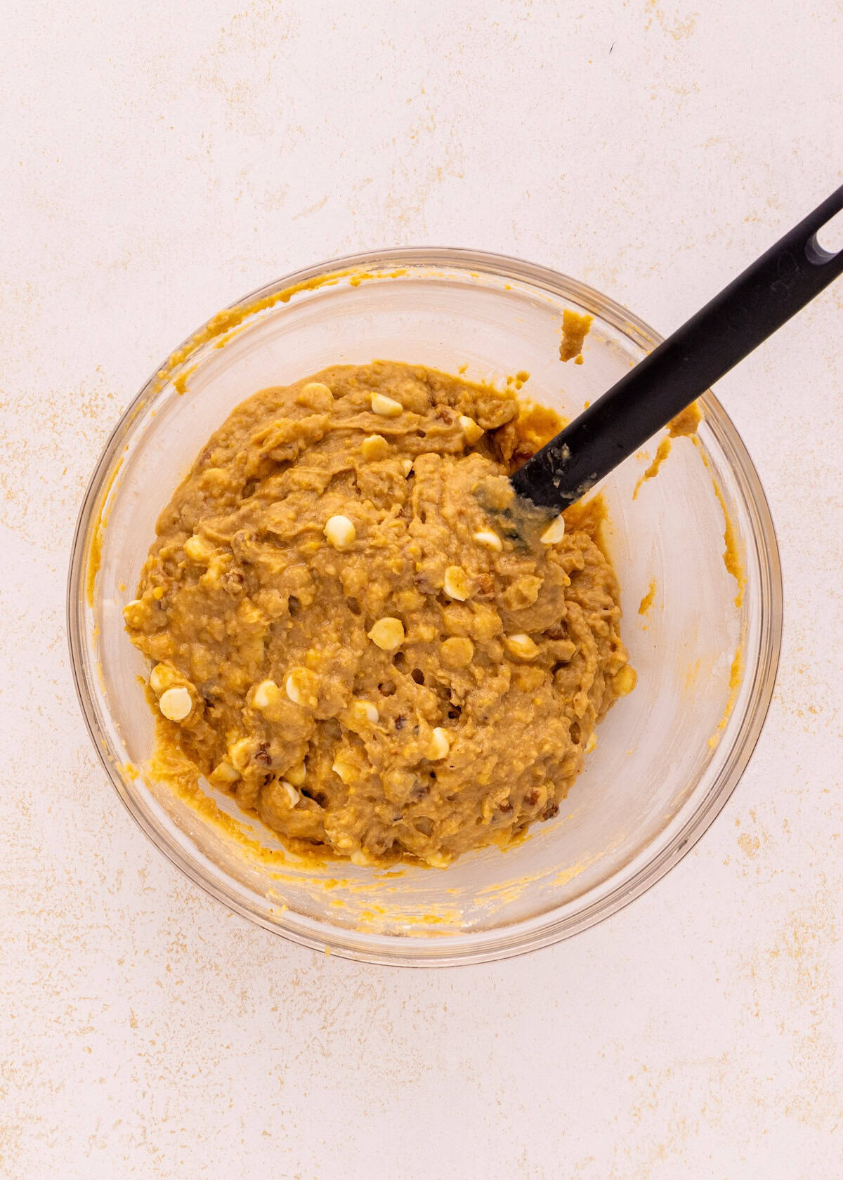 Bowl of cookie dough with white chocolate chips and a black spatula on a light surface.