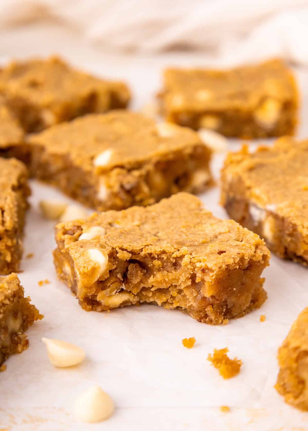 Blondie bars with white chocolate chips, one with a bite taken out, on parchment paper.