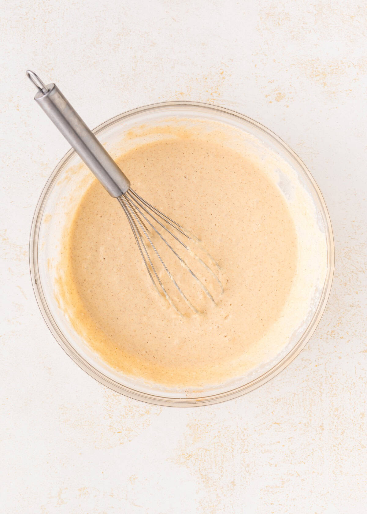 A glass bowl of pancake batter with a metal whisk on a light surface.