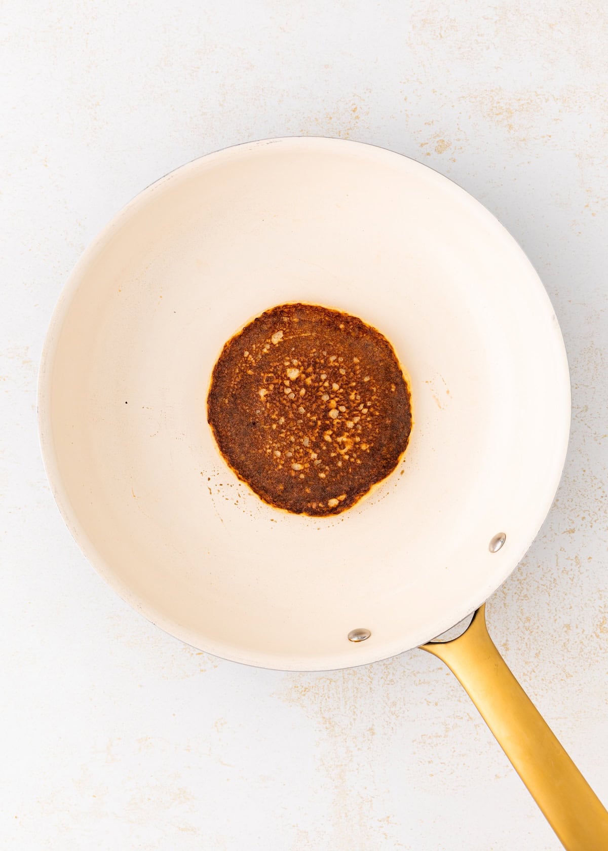 A single burnt pancake sits in the center of a white frying pan with a gold handle.