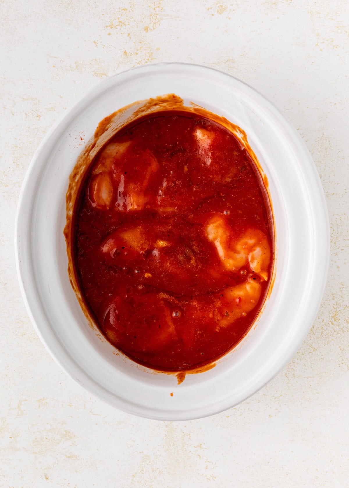 Raw chicken breasts covered in red sauce inside a white oval slow cooker on a light background.