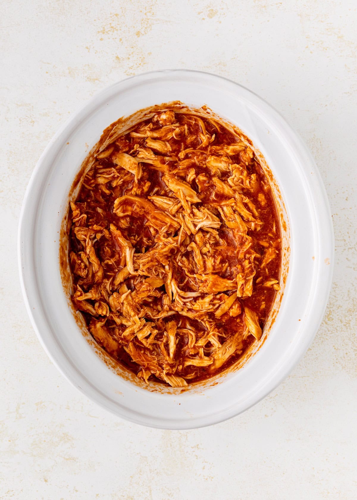 Shredded barbecue chicken in a white oval slow cooker on a light background.