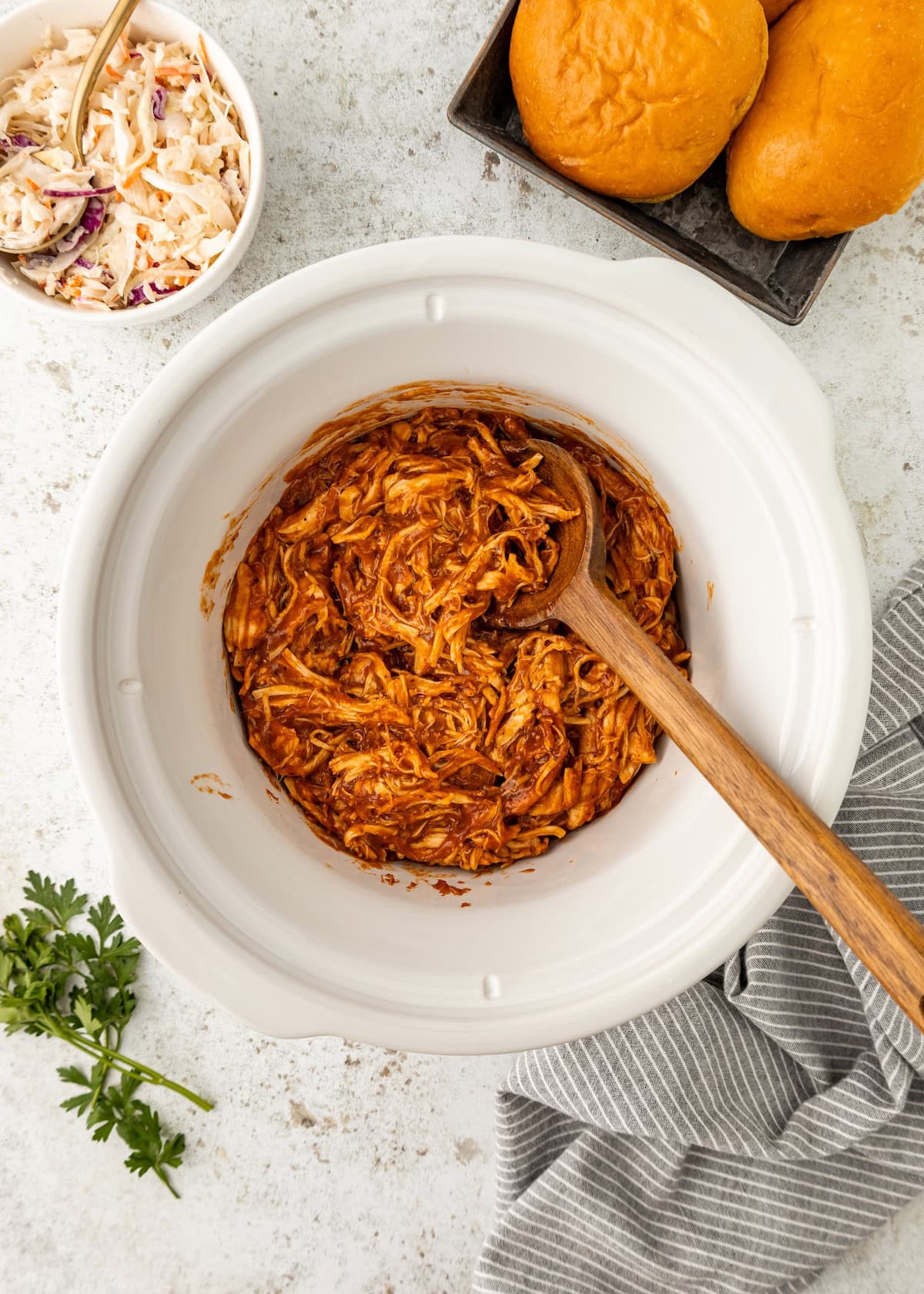 Shredded barbecue chicken in a slow cooker with a wooden spoon, buns, and coleslaw nearby.